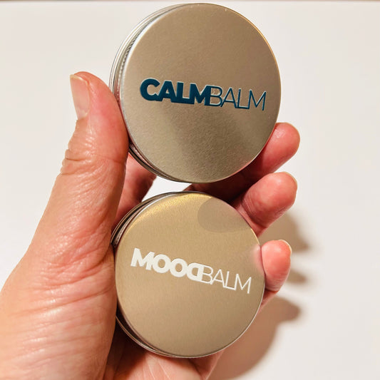 A hand holding two round metal tins of mood balm, one labeled CALM BALM in blue and the other labeled MOOD BALM in white, against a plain light background.