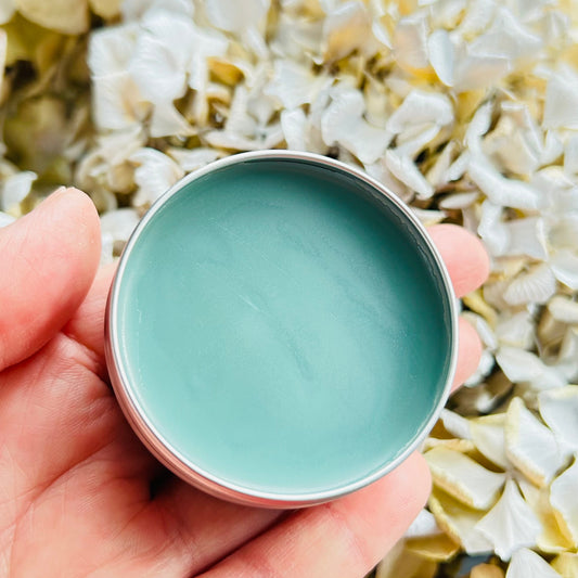 A hand holding a small round metal tin filled with smooth, pale mint-green balm or cream, set against a soft background of white and pale yellow dried flowers.