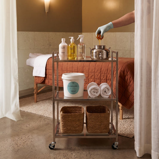 A metal three-tier rolling cart stocked with spa products, towels, and baskets, positioned in front of a massage table in a warm-toned treatment room. A gloved hand is pouring honey into a metal pot on the cart.