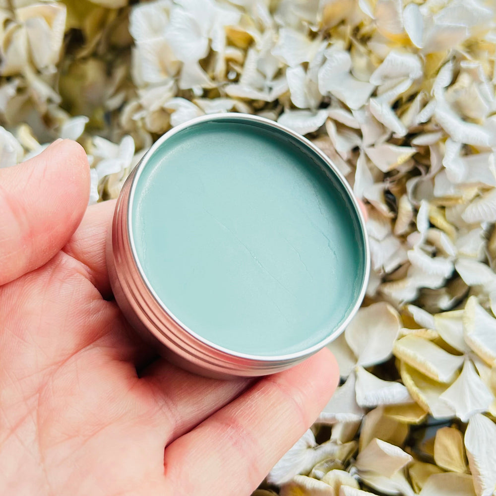 Hand holding a small container of light blue balm against a floral background