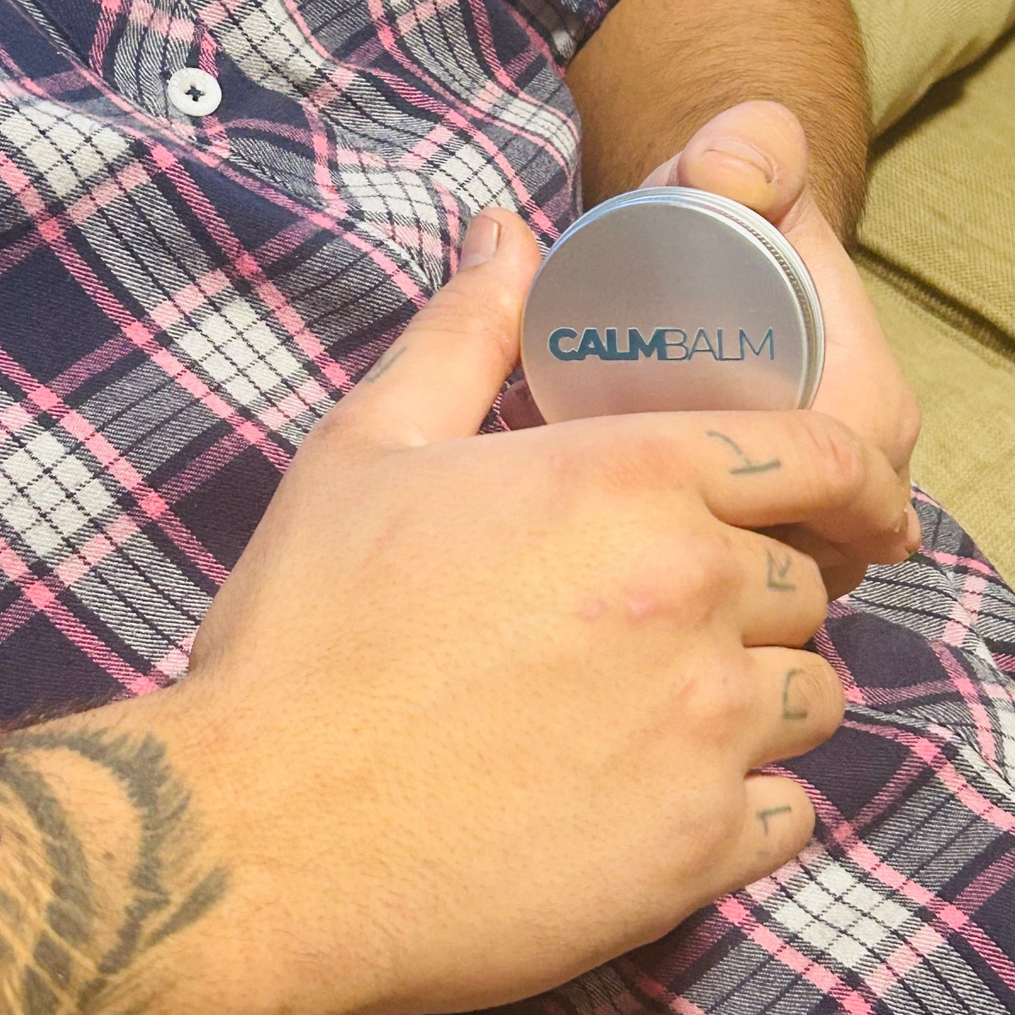 A close-up of a person holding a round silver tin labeled 'CALM BALM' against their chest, with a tattooed hand resting over it. The person is wearing a navy, pink, and white plaid shirt.