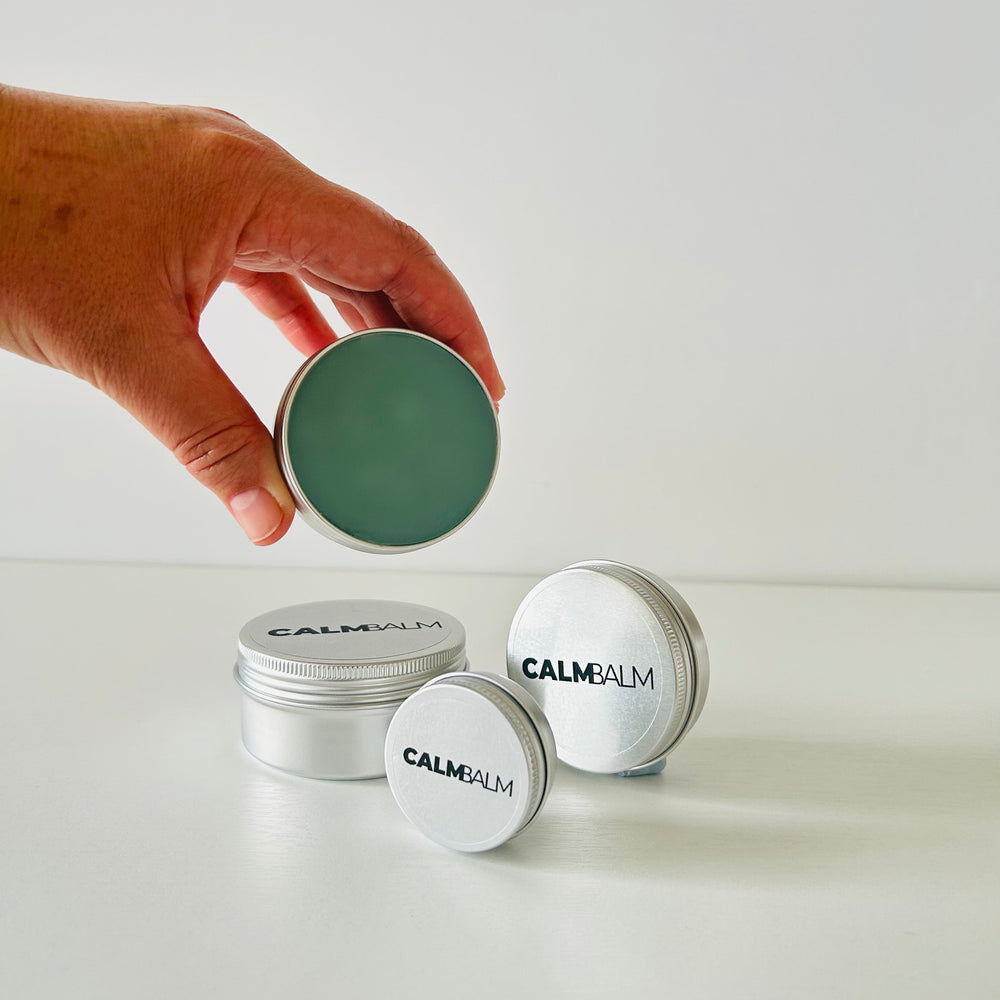 Hand opening a container of Calm Balm with additional containers stacked below on a white background.