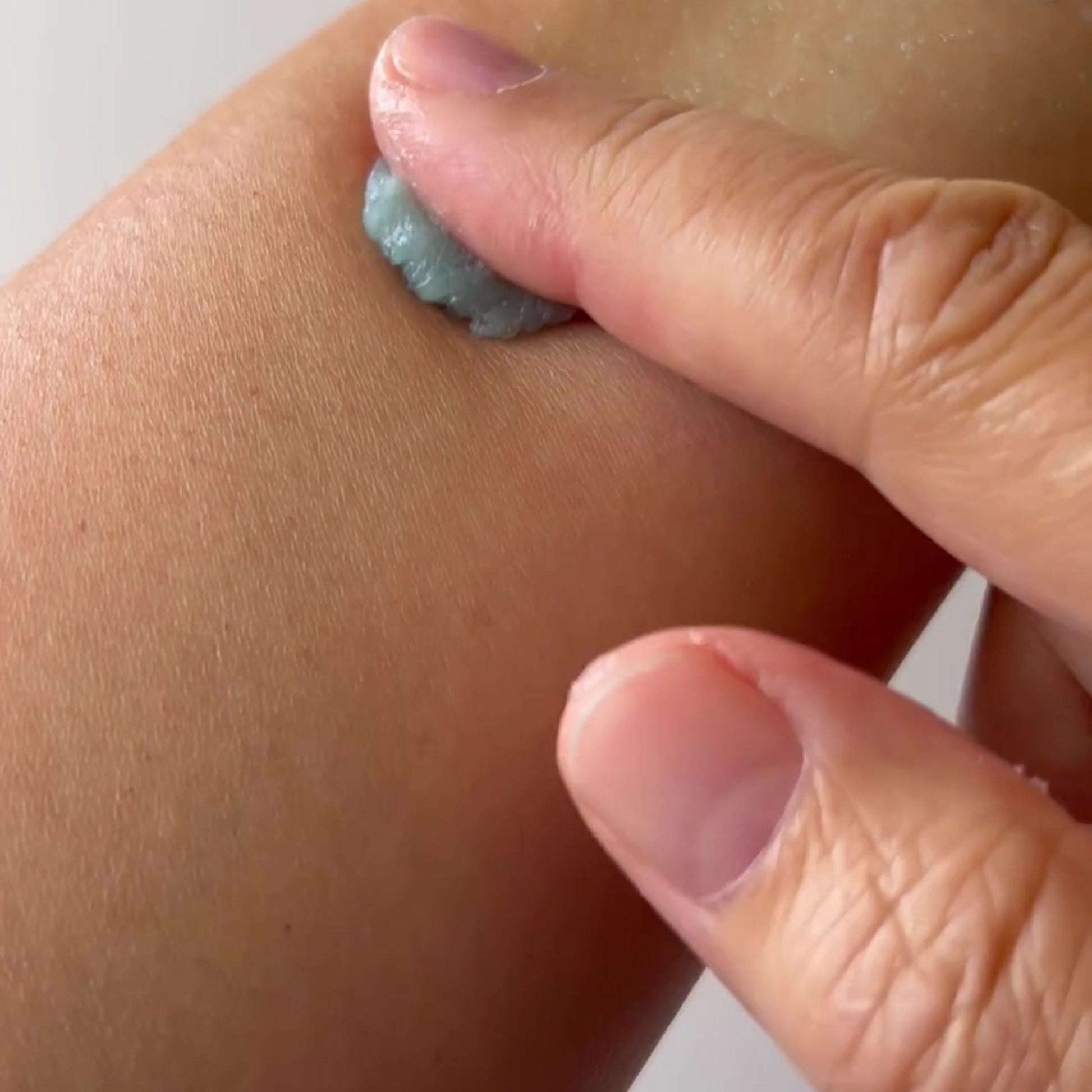 A close-up of a person's hand applying a thick, pale blue-gray scrub or cream onto their forearm.