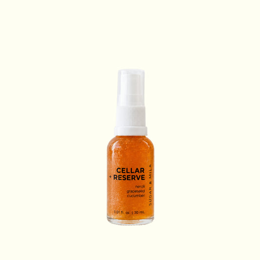 A small glass spray bottle of Cellar Reserve Neroli Green Sandalwood Cucumber body spray, filled with orange liquid and suspended in fine gold glitter, set against a solid cream background.