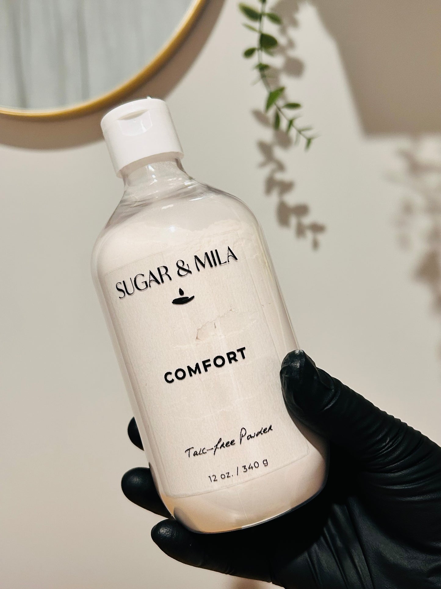 A hand wearing a black glove holds a clear plastic bottle of Sugar & Mila Comfort Talc-Free Powder against a warm beige wall with soft shadows and a hanging green vine.