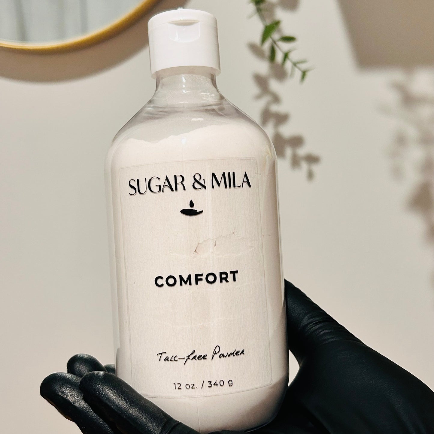 A hand wearing a black glove holds a clear plastic bottle of Sugar & Mila Comfort Talc-Free Powder. The bottle is frosted, showing the white powder inside, and is set against a warm beige wall with soft shadows and a hint of a mirror in the background.