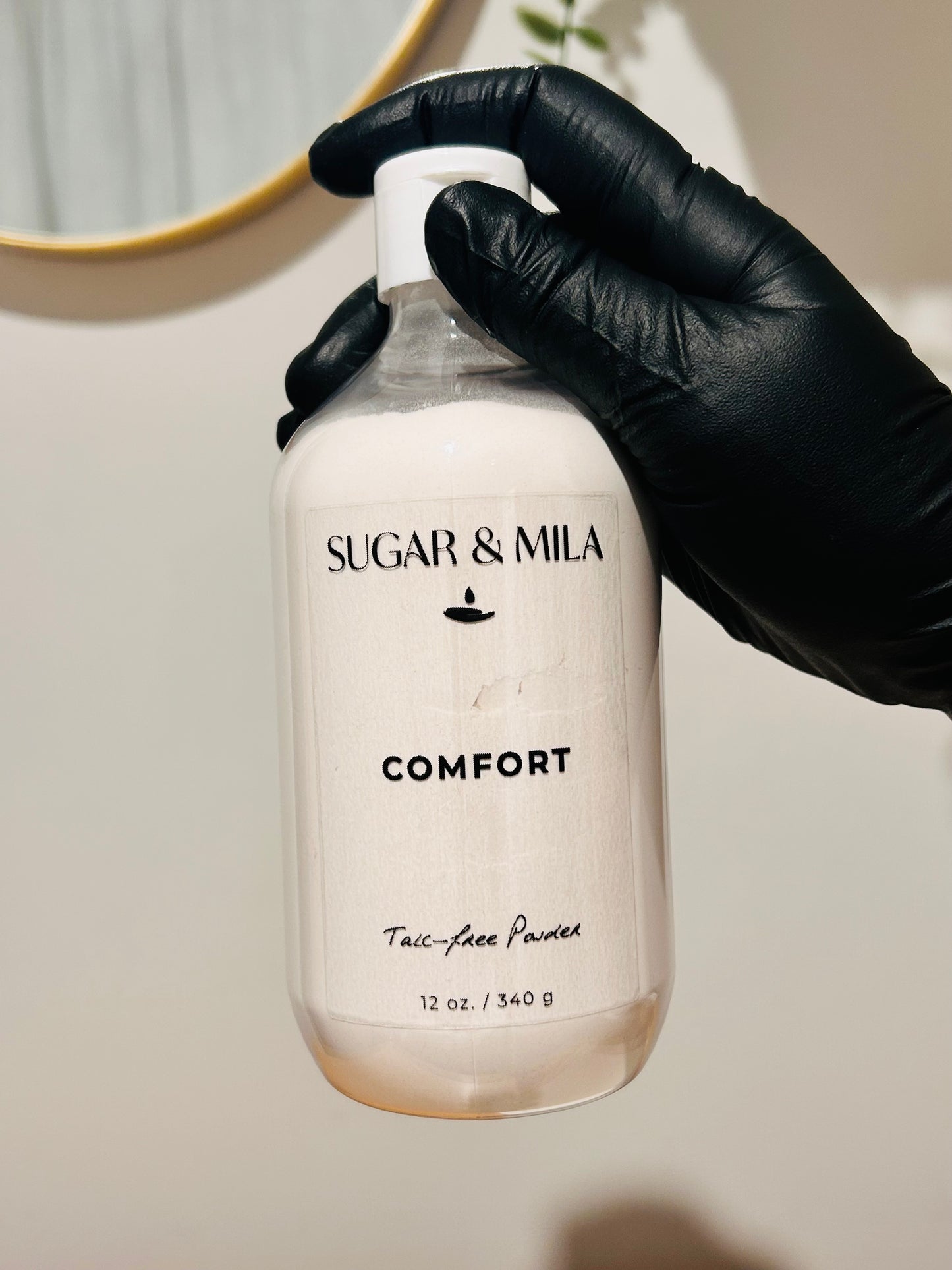 A hand wearing a black glove holds a clear plastic bottle of Sugar & Mila Comfort Talc-Face Powder against a warm beige background.