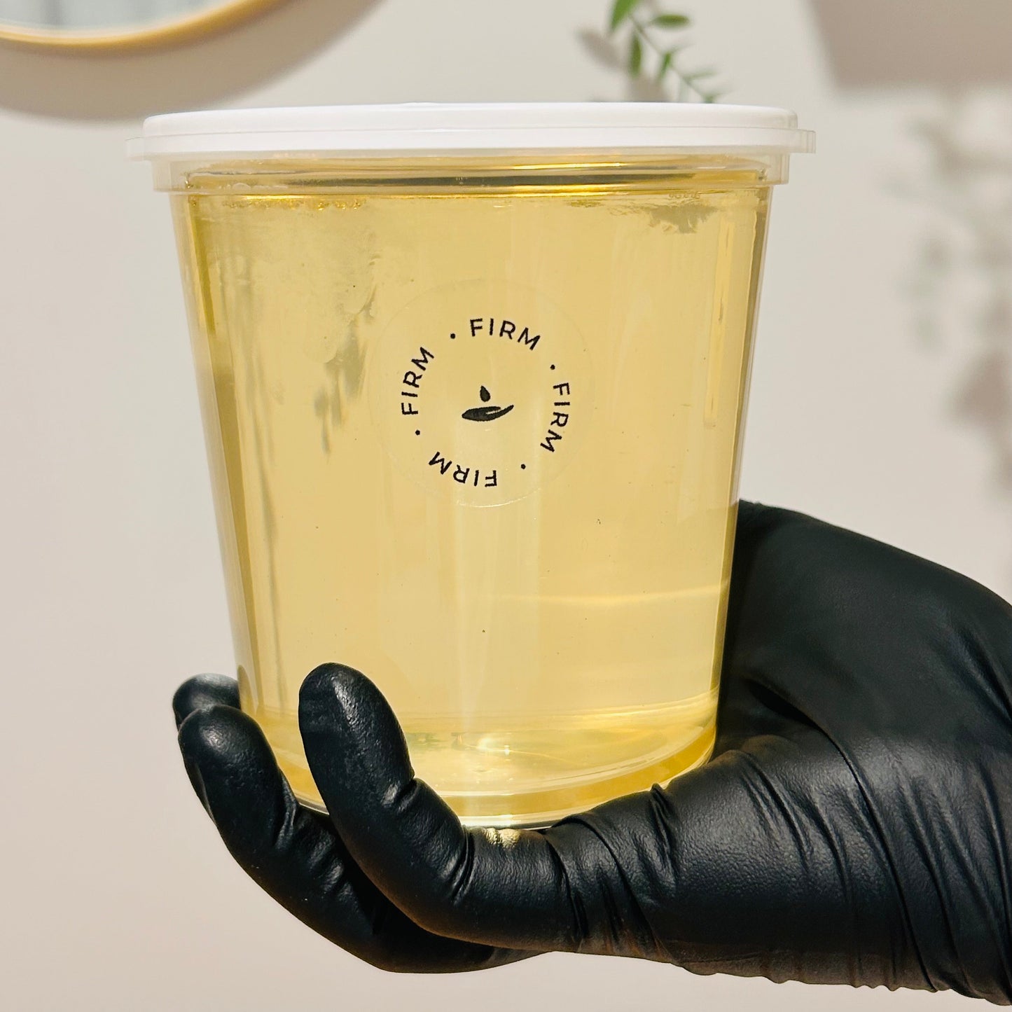 A hand wearing a black glove holds a clear plastic tub filled with pale yellow gel or liquid, featuring a white lid and a black circular logo that reads 'FIRM' around a small droplet icon. The background is a softly lit indoor wall with a mirror edge and greenery visible.