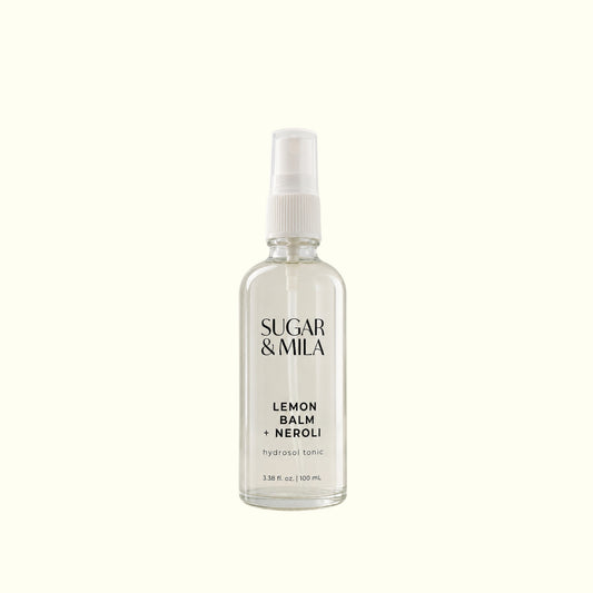 A clear glass spray bottle of Sugar & Mila Lemon Balm + Neroli body spray with a white pump dispenser, centered on a solid cream background.