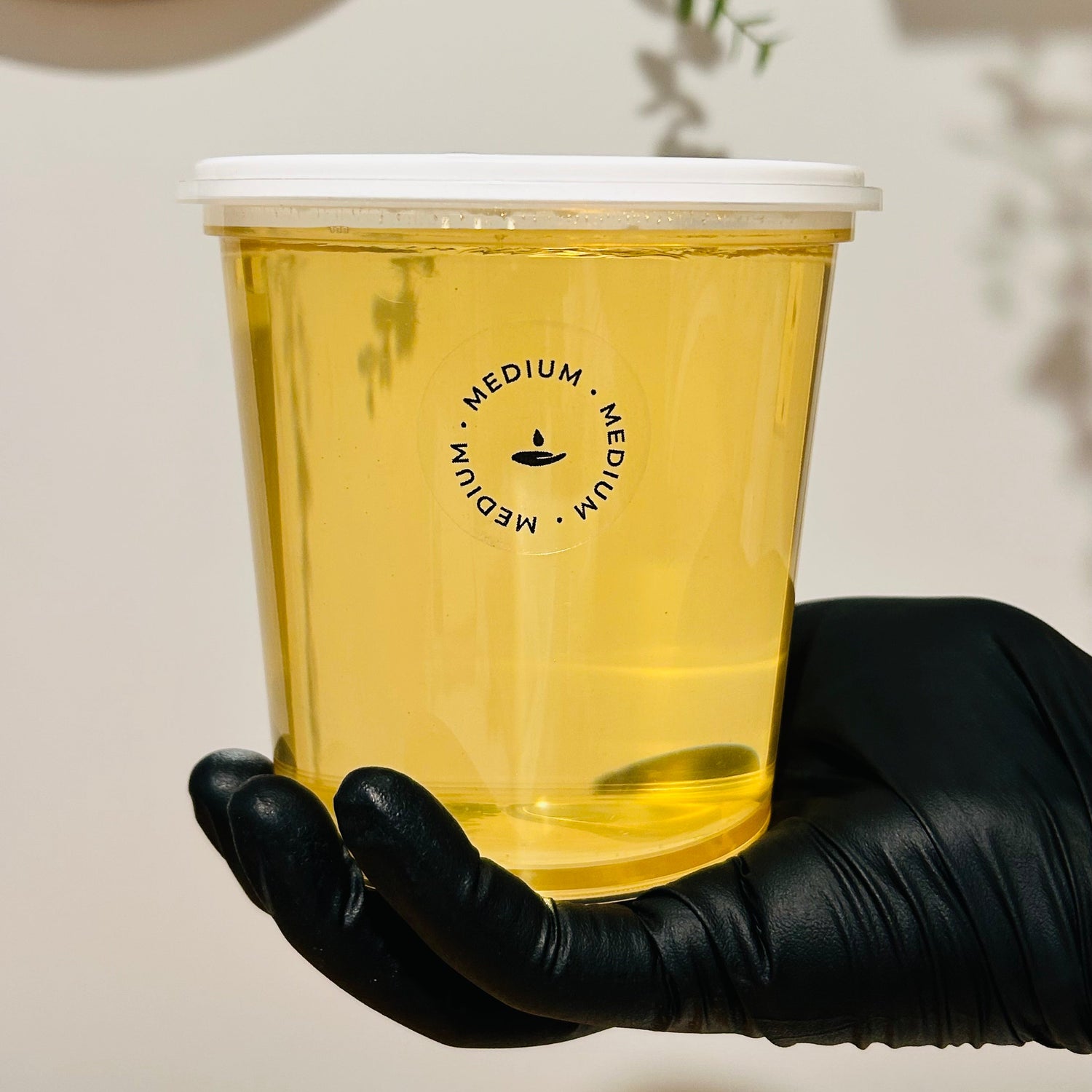 A clear plastic cup filled with golden yellow liquid, held by a hand wearing a black glove. The cup has a white lid and a black circular logo that reads 'MEDIUM'.