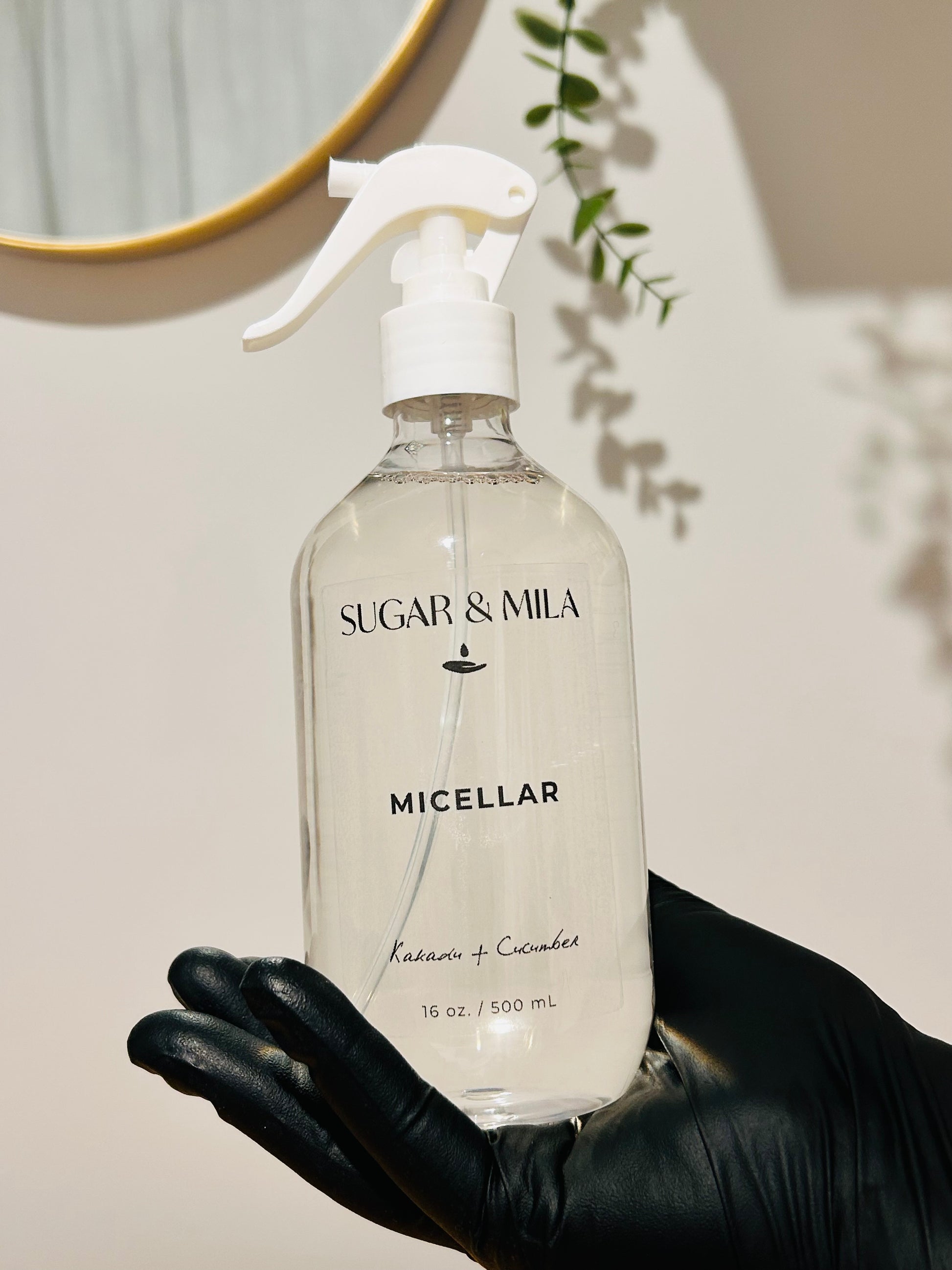 A clear plastic spray bottle of Sugar & Mila Micellar Cleanser with a white trigger sprayer, held in a hand wearing a black glove against a warm beige wall with soft shadows.