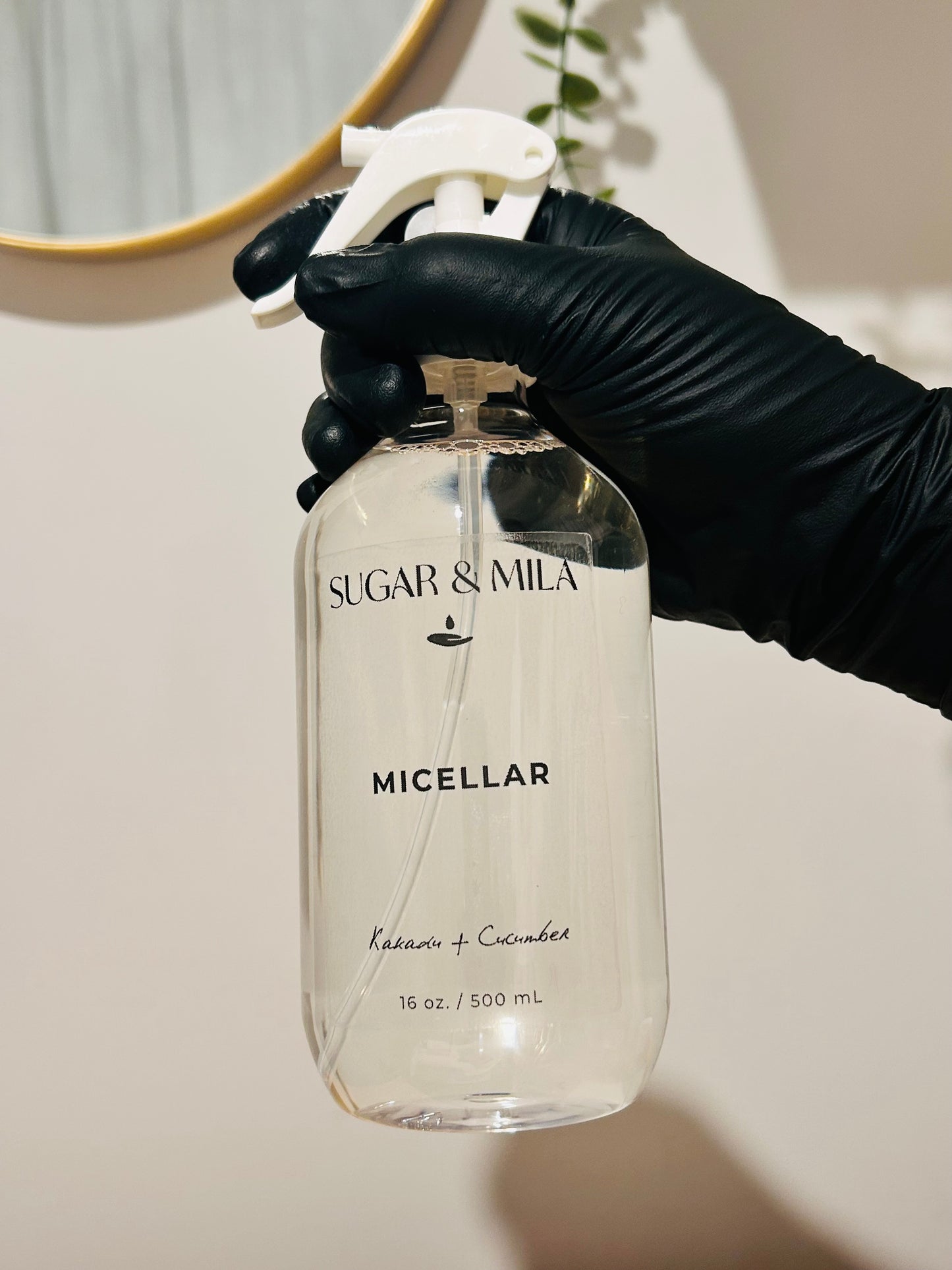 A hand wearing a black glove holds a clear plastic spray bottle of Sugar & Mila Micellar cleanser against a neutral beige wall background.