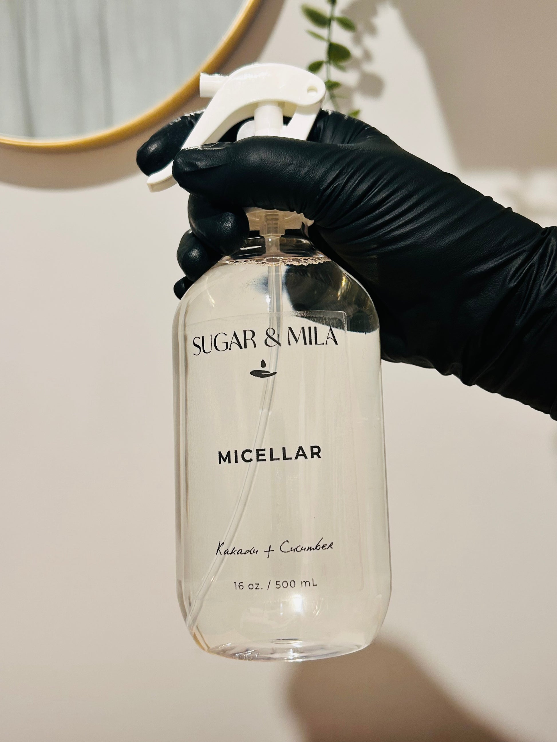 A hand wearing a black glove holds a clear plastic spray bottle of Sugar & Mila Micellar cleanser against a neutral beige wall background.