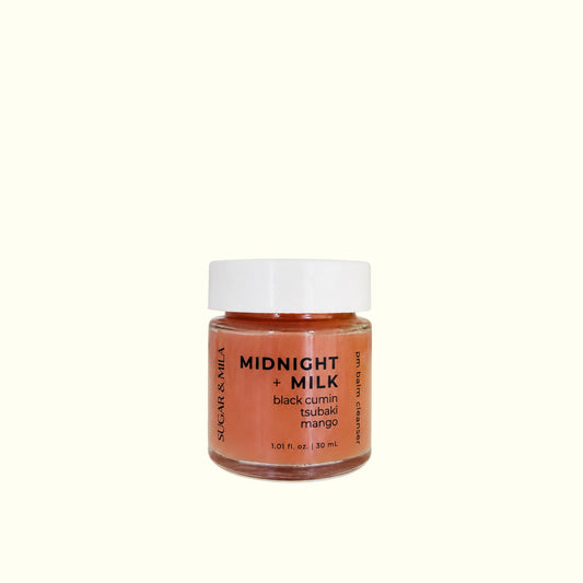 A small glass jar of St. Gar & Nila 'Midnight + Milk' black cumin tsubaki mango tsogat, featuring a white lid and a label with black and orange text, set against a solid off-white background.