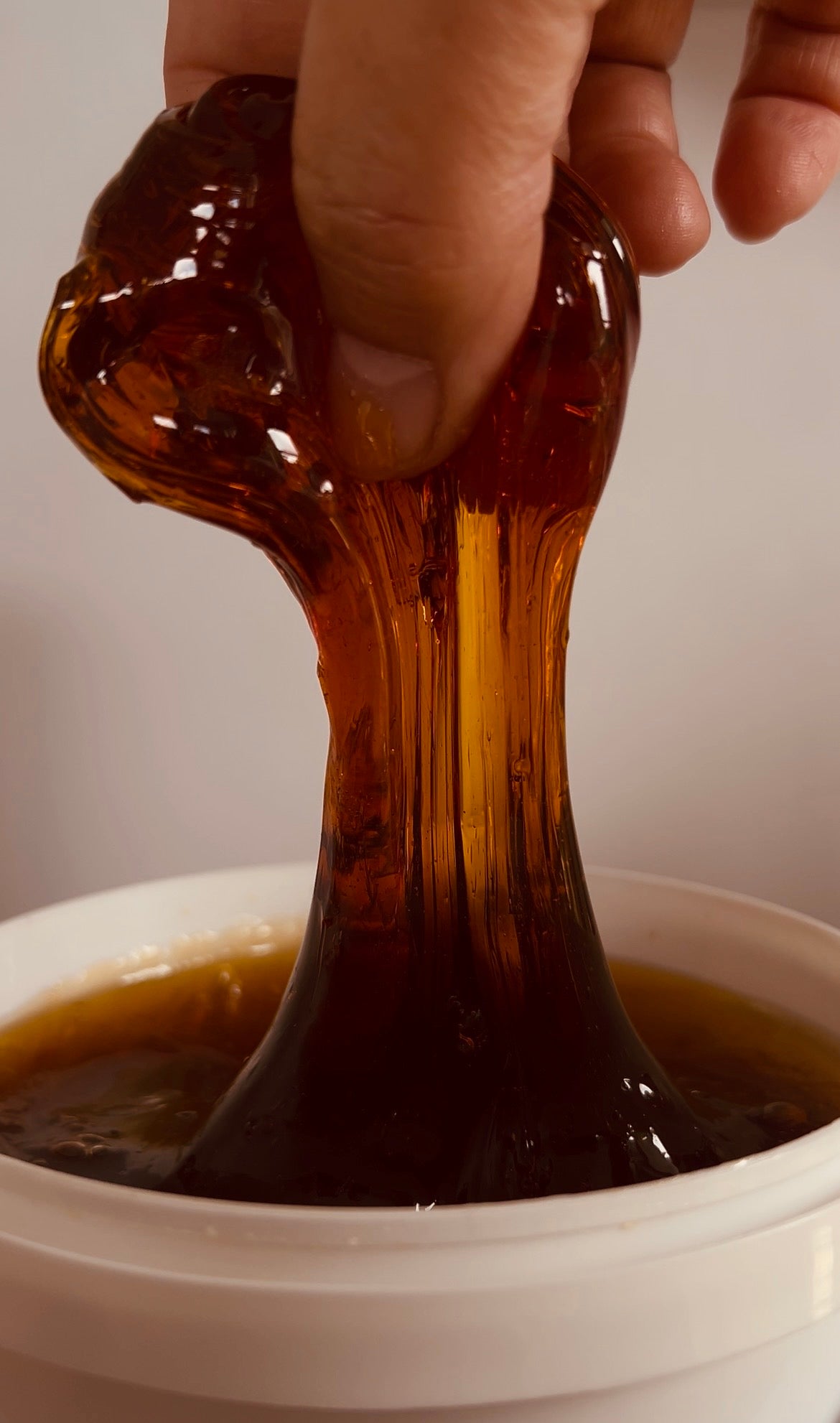 A hand stretching a glossy, amber-colored translucent slime or resin above a white bowl filled with a darker brown liquid.