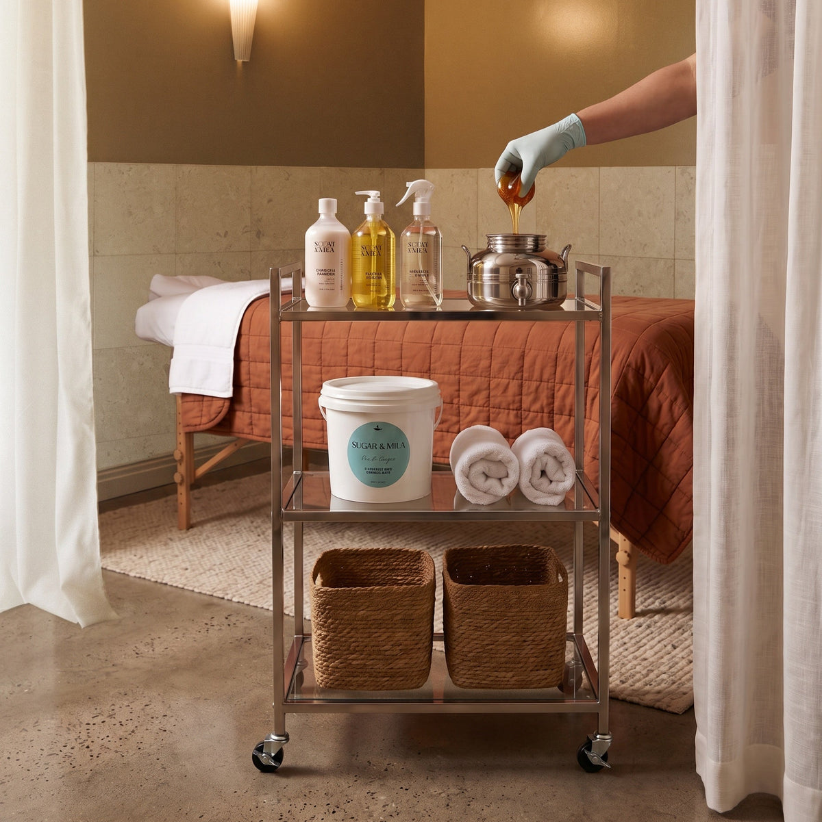 A metal three-tier rolling cart stocked with spa products, towels, and baskets, positioned in front of a massage table in a warm-toned treatment room. A gloved hand is pouring honey into a metal pot on the cart.