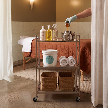 A metal three-tier rolling cart stocked with spa products, towels, and baskets, positioned in front of a massage table in a warm-toned treatment room. A gloved hand is pouring honey into a metal pot on the cart.