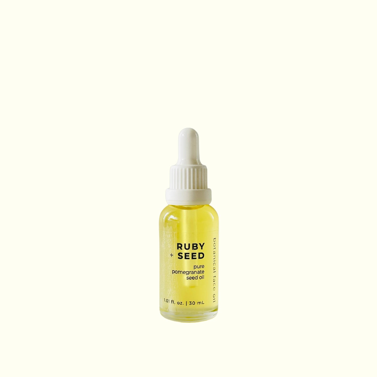 A small glass dropper bottle of Ruby + Seed pure pomegranate seed oil with a white cap, centered on a solid cream background.