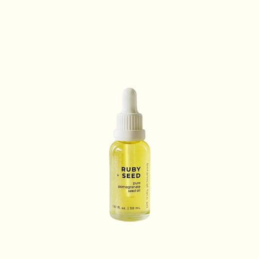 A small glass dropper bottle of Ruby + Seed pure pomegranate seed oil with a white cap, centered on a solid cream background.