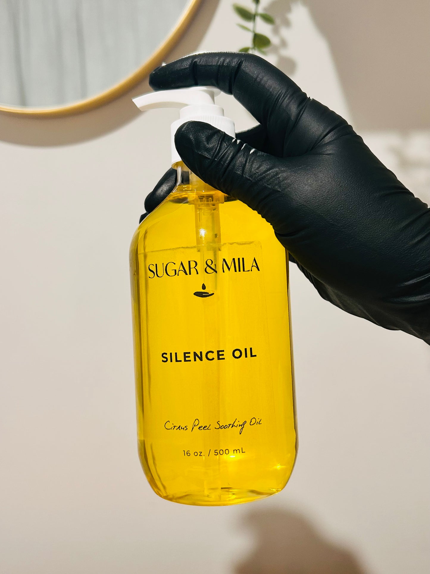 A hand wearing a black glove holds a clear plastic pump bottle filled with bright yellow oil. The bottle is labeled 'SUGAR & MILA SILENCE OIL' and is photographed against a light beige wall with a gold-framed mirror partially visible in the background.