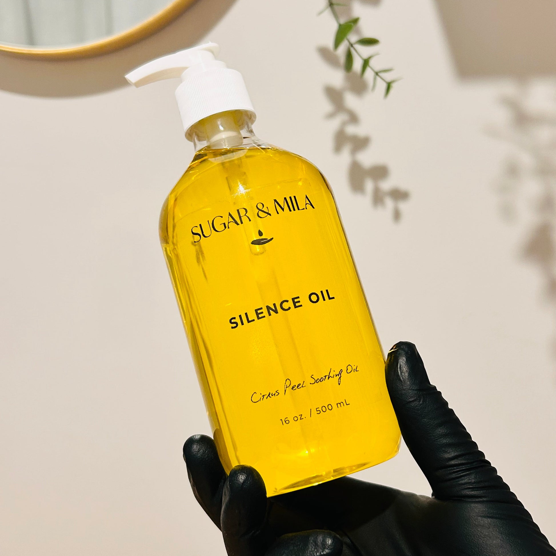 A hand wearing a black glove holds a clear bottle of Sugar & Mila Silence Oil, showing the bright yellow oil inside, against a warm beige wall with soft shadows.