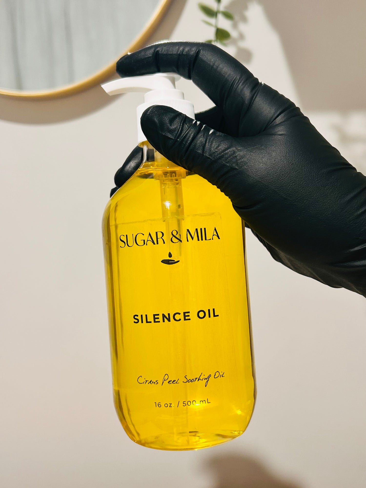 A hand wearing a black glove holds a clear plastic pump bottle filled with bright yellow oil. The bottle is labeled 'SUGAR & MILA SILENCE OIL' and is photographed against a warm beige wall with a softly blurred mirror and plant in the background.