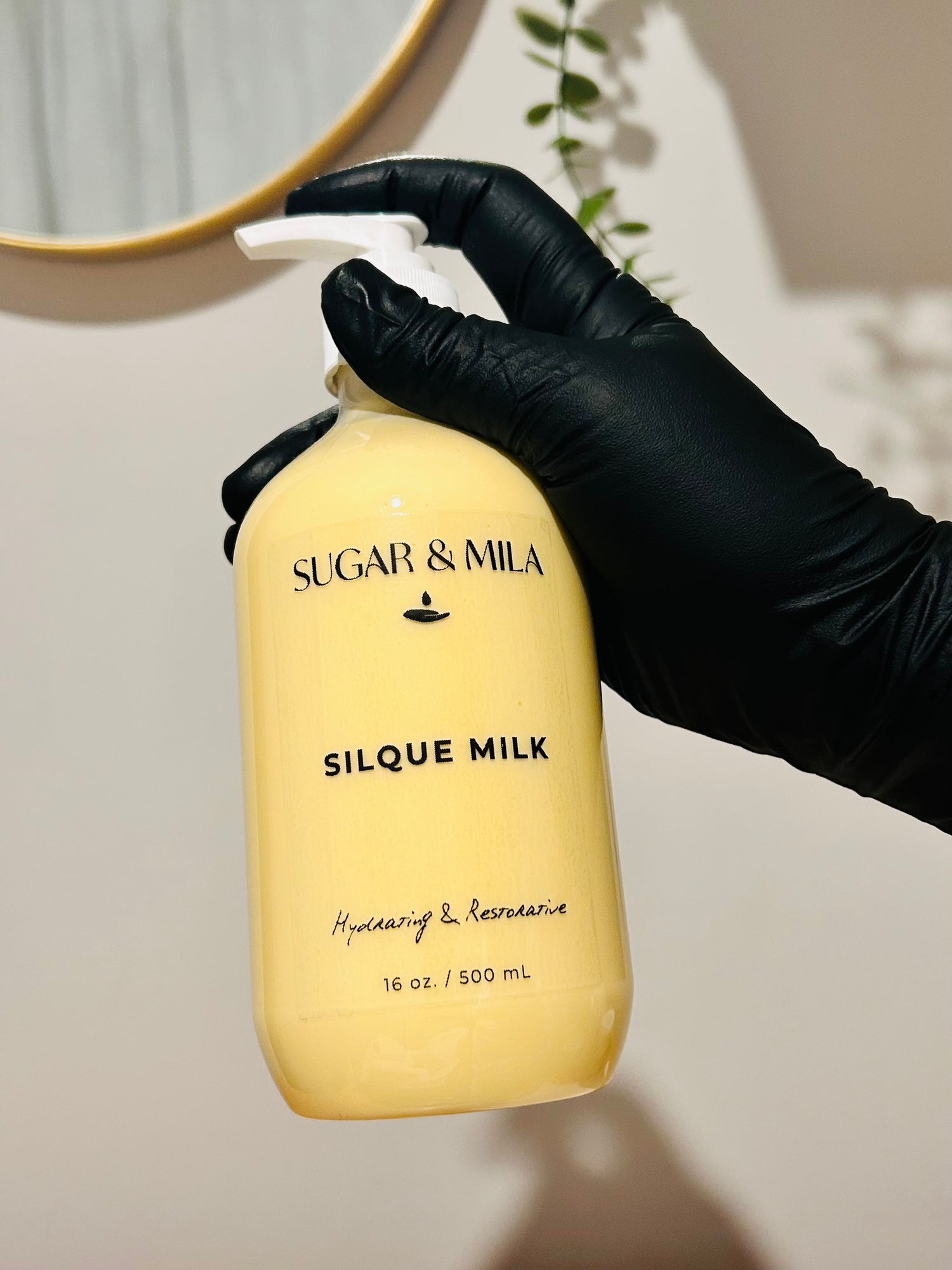 A hand wearing a black glove holds a pump bottle of Sugar & Mila Silque Milk Hydrating & Rejuvenating serum against a neutral indoor background.