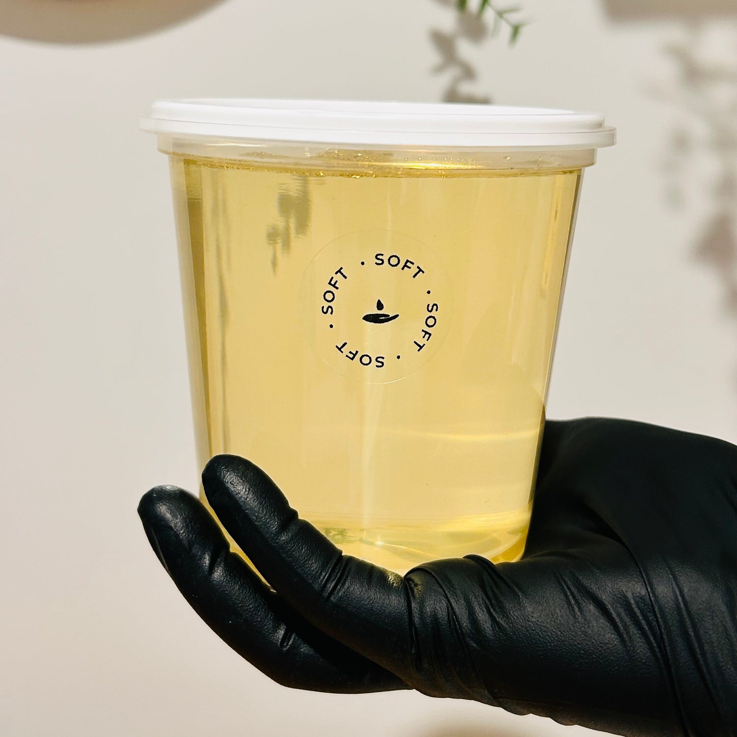 A hand wearing a black glove holds a clear plastic cup filled with pale yellow liquid. The cup has a white lid and a black circular logo reading 'SOFT • SOFT • SOFT' with a small lamp icon.