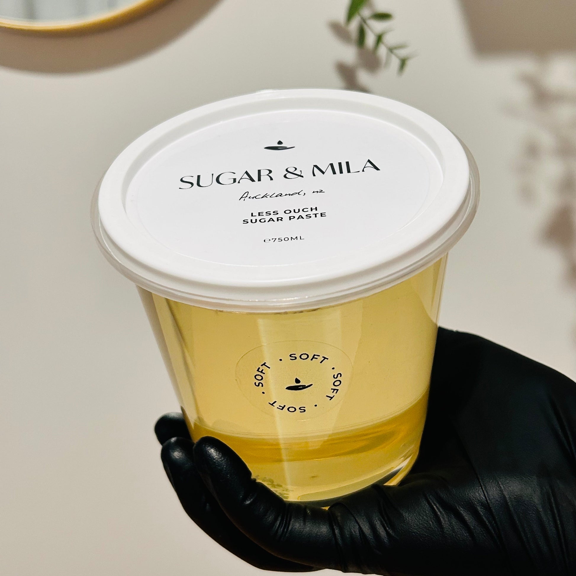 A hand wearing a black glove holds a clear plastic jar of Sugar & Mila free sugar sugar paste, showing the pale yellow paste inside. The product has a white lid with black text, set against a warm beige background with a gold-framed mirror and a green plant in the distance.