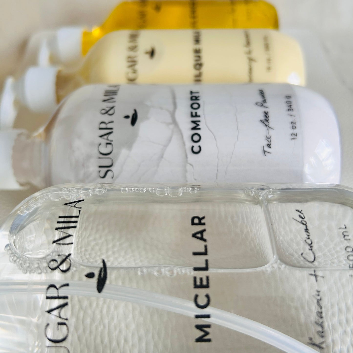 Four bottles of Sugar & Mila skincare products lined up diagonally, including a micellar cleanser, a powder, and lotion in clear packaging.