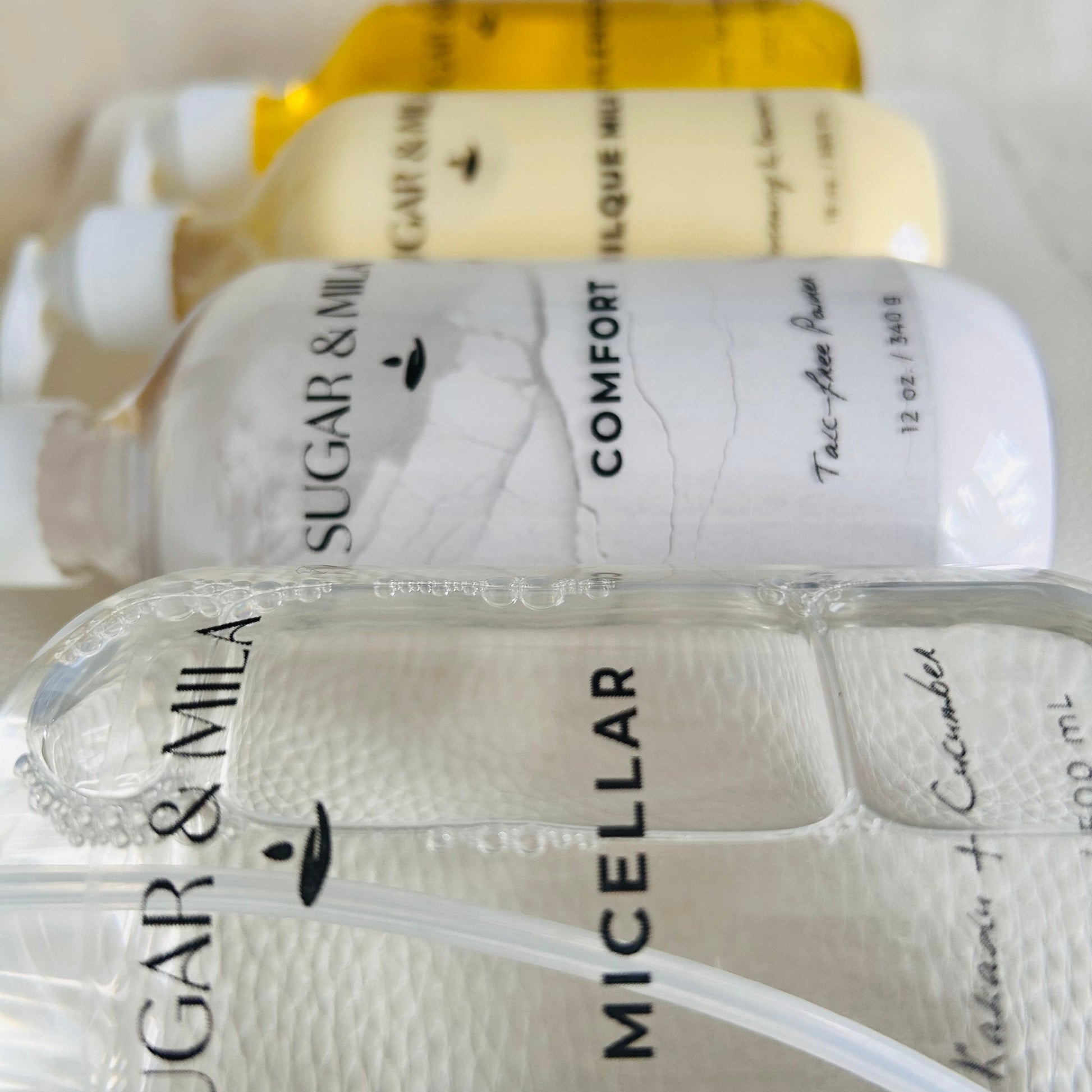 Four bottles of Sugar & Mila skincare products lined up diagonally, including a micellar cleanser, a powder, and lotion in clear packaging.