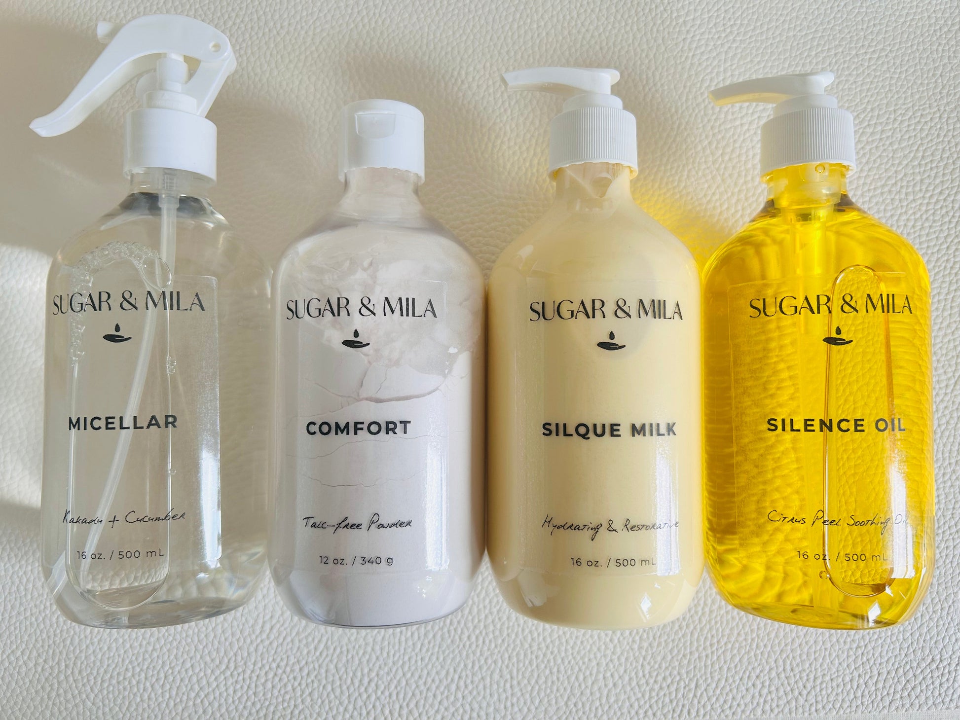 Four Sugar & Mila skincare bottles lined up against a textured white wall: Micellar, Comfort, Silque Milk, and Silence Oil.