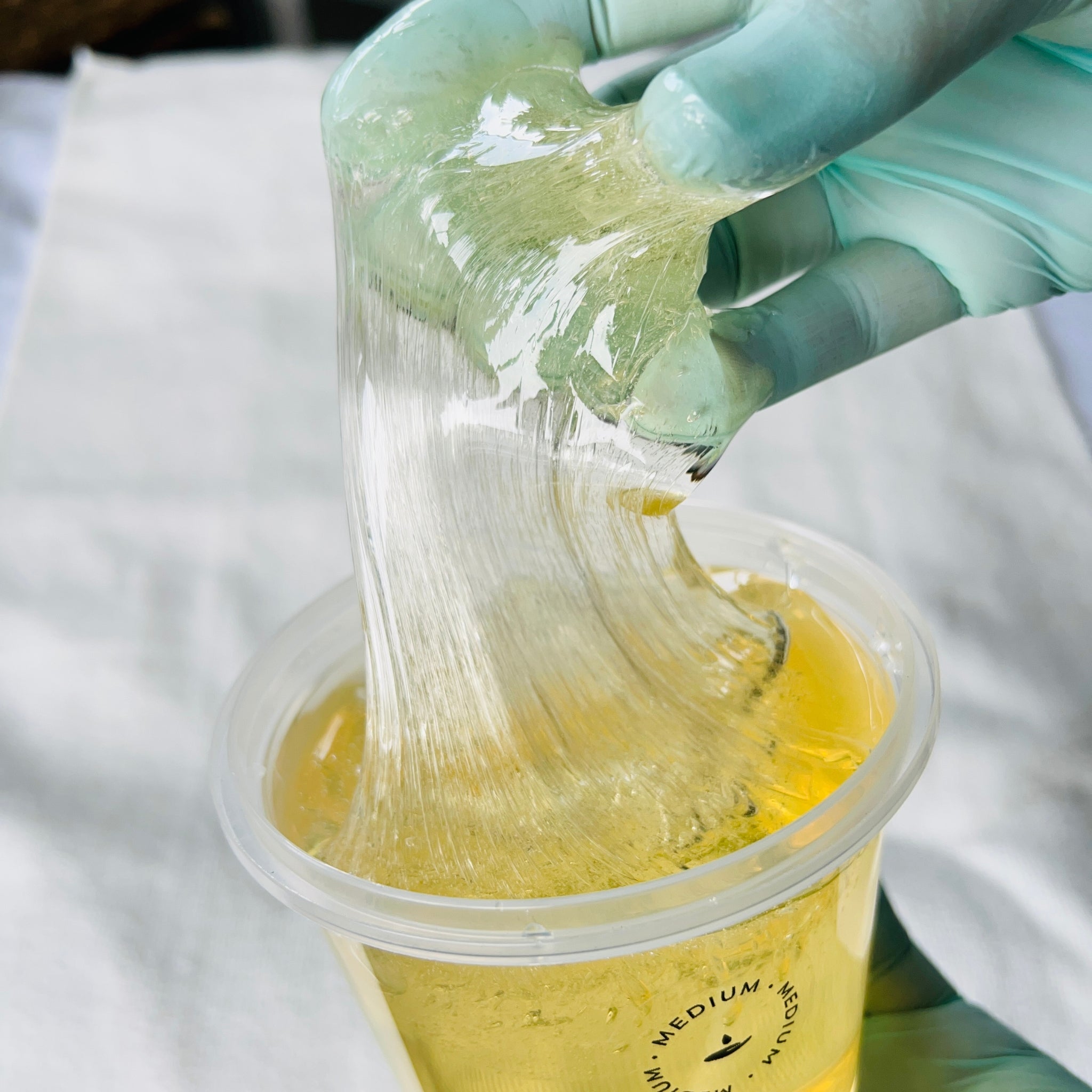 Hand wearing a blue glove stretching a yellowish substance over a container labeled 'Medium'.