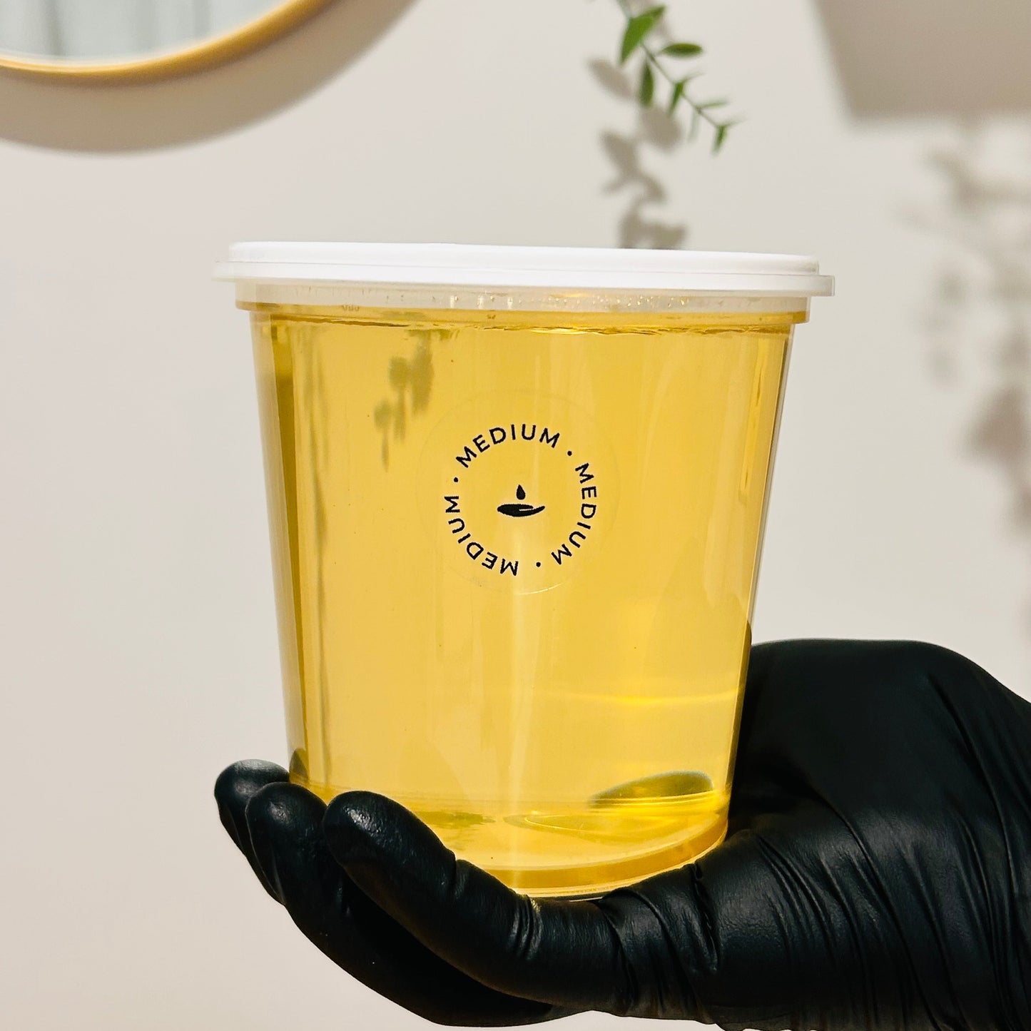 A hand wearing a black glove holds a translucent yellow-tinted plastic cup with a white lid. The cup has a black circular logo reading 'MEDIUM' with a small symbol in the center.