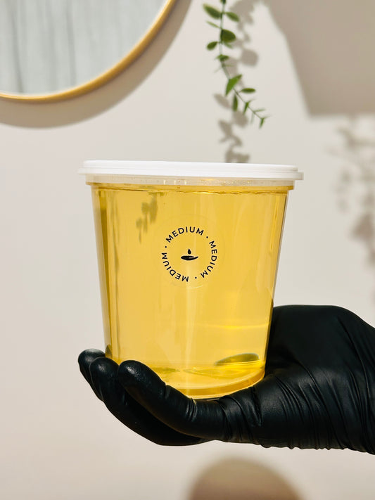 A hand wearing a black glove holds a translucent yellow-tinted plastic cup with a white lid. The cup has a black circular logo reading 'MEDIUM' with a small symbol in the center.